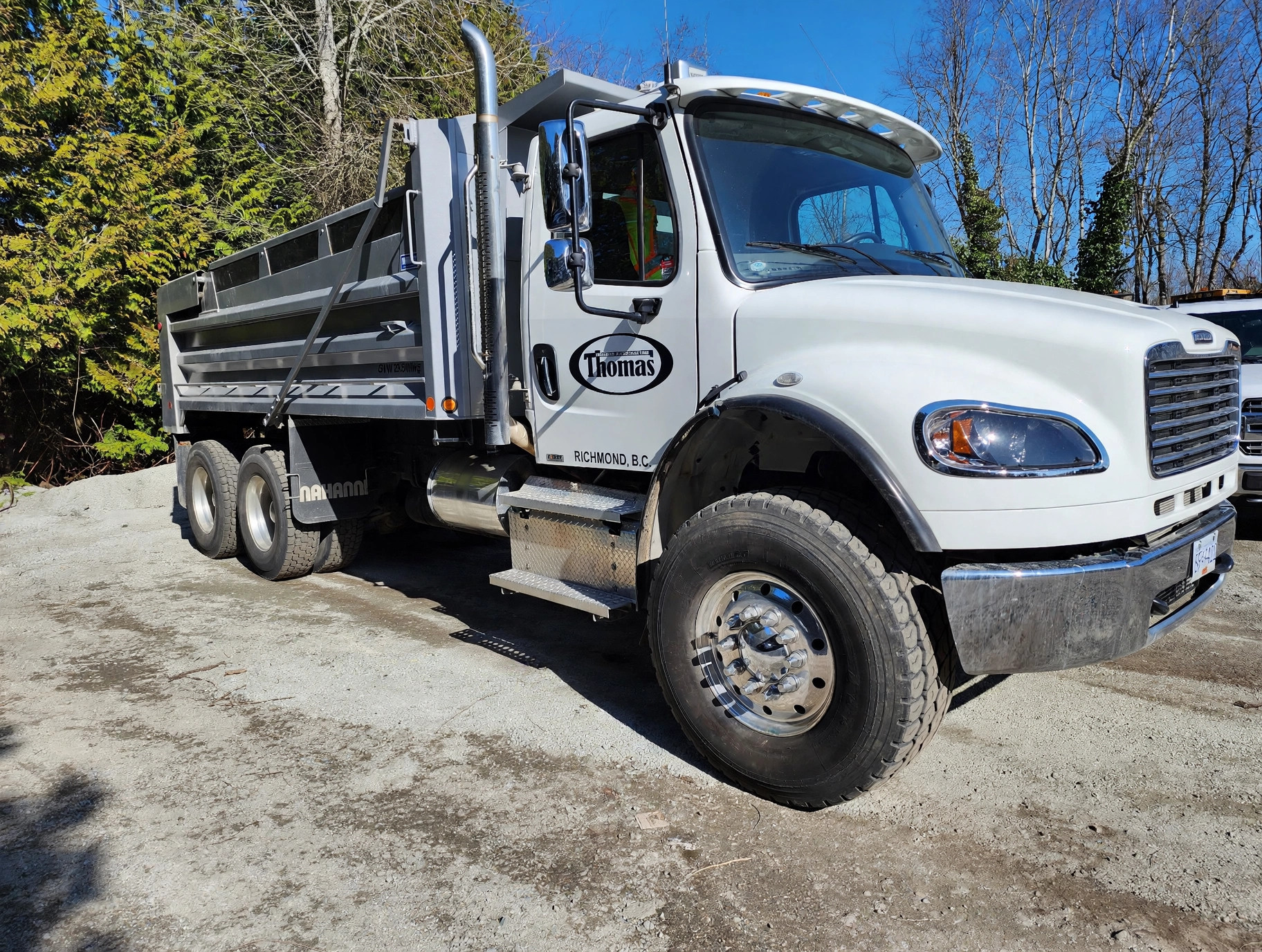 White Thomas Trucking dump truck with the Thomas oval logo and Richmond BC lettering on the door, parked on gravel with trees behind