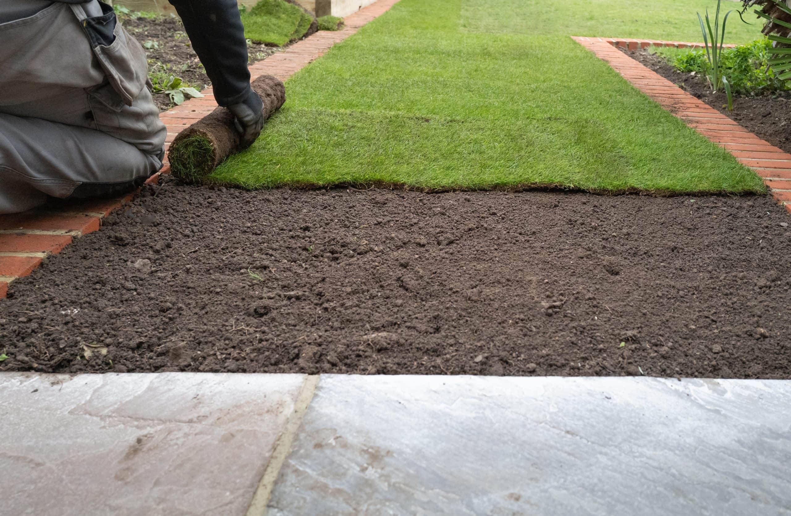 Fresh green turf being unrolled over prepared dark topsoil in a neat garden bordered by brick edging